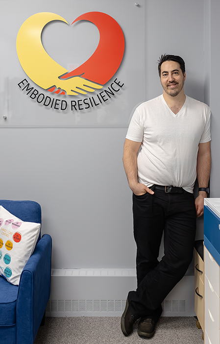 Robert Simms standing in a corner with an Embodied Resilience logo plaque above his right shoulder.